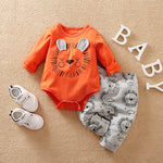 Orange baby onesie with lion design, gray pants with lion patterns, and white baby shoes on a beige surface.
