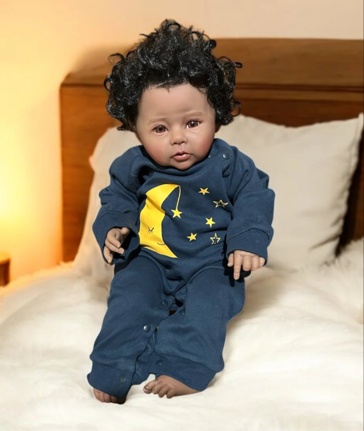 A charming baby doll rests on a bed, with a phone nearby, wearing a unisex nighty night jumpsuit.