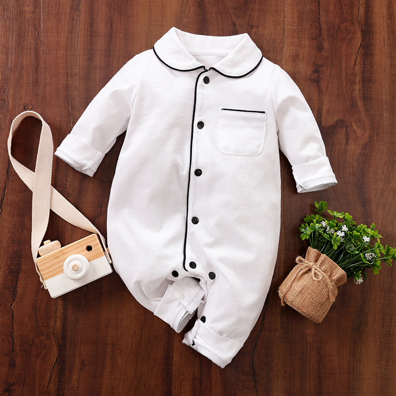 White baby cotton pajamas with black snap buttons on a wooden surface, accompanied by a toy camera and small plant.