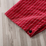 A close-up of the hem of the Adorable 1-Piece Striped Infant Clothing Jumpsuit With Bow Tie & Button-Up Front, showing red fabric with thin white vertical stripes and a black button, lying on a light wood-grain floor.
