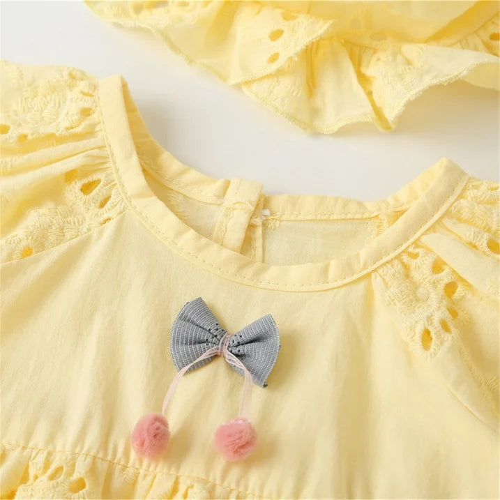 Soft yellow baby dress featuring eyelet sleeves and a grey bow with pink pom-poms.