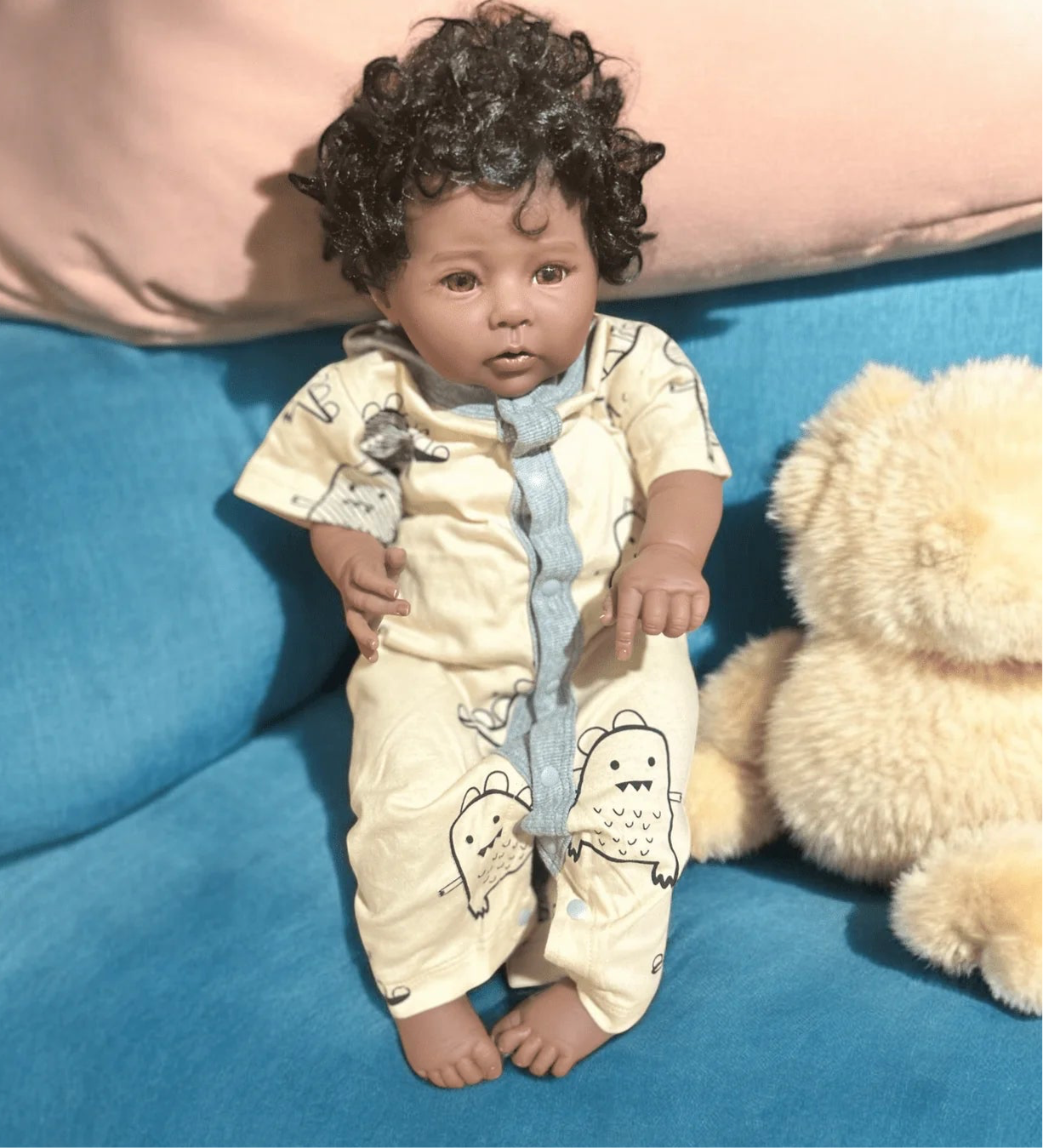 A baby doll rests on a couch with a teddy bear, featuring a delightful unisex baby romper perfect for any little one.