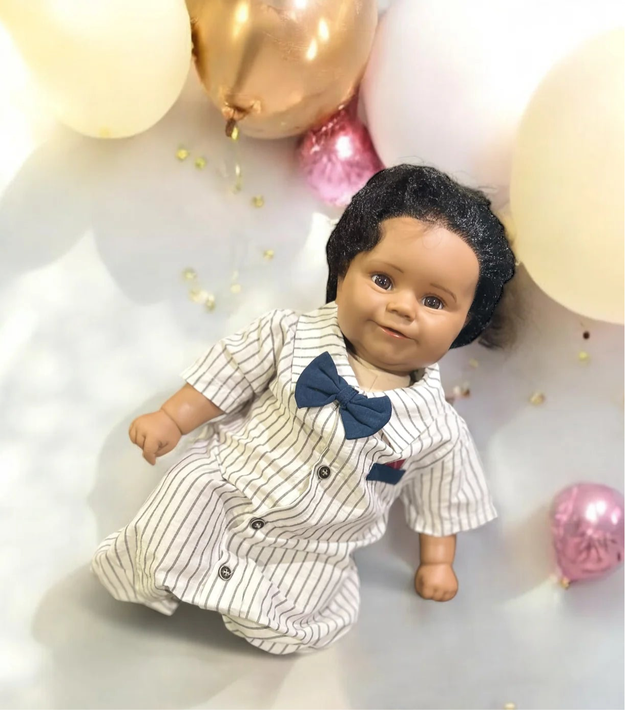 An adorable baby doll dressed in a striped jumpsuit and bow tie, peacefully lying on a table, ready for playtime.