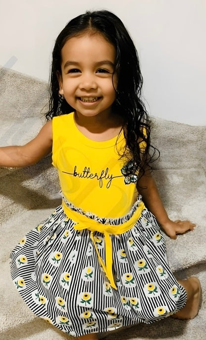 A cheerful girl in a yellow dress paired with a striped shirt, embodying playful spirit and style for toddlers.