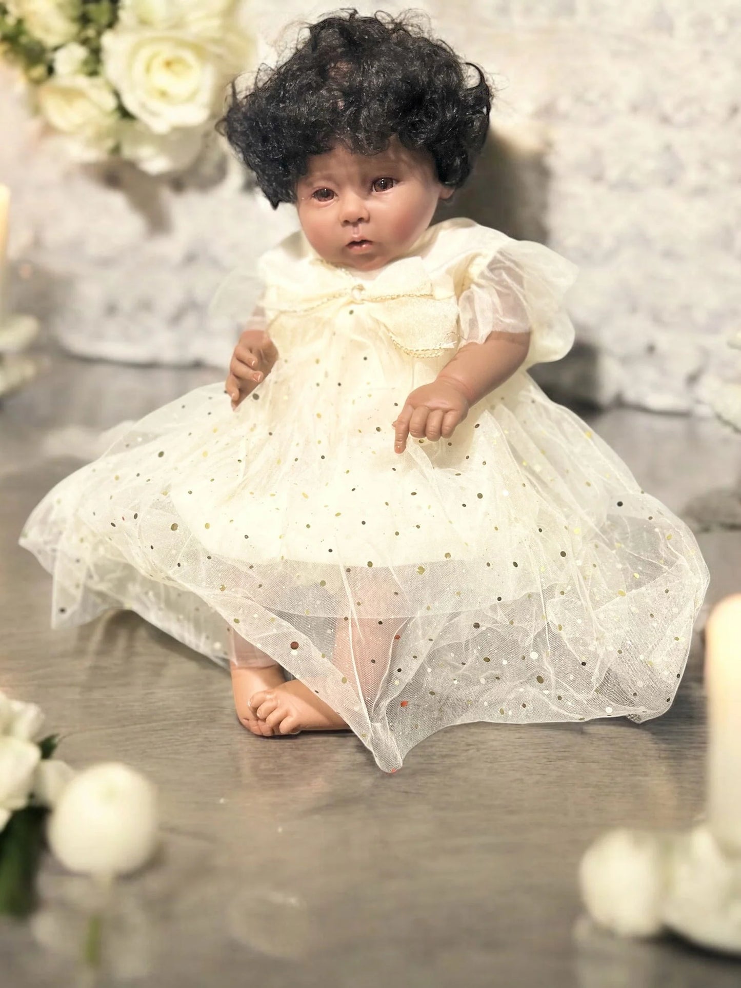 A delightful baby doll wearing a sparkling ivory romper dress sits on a table, embodying sweetness and playfulness.